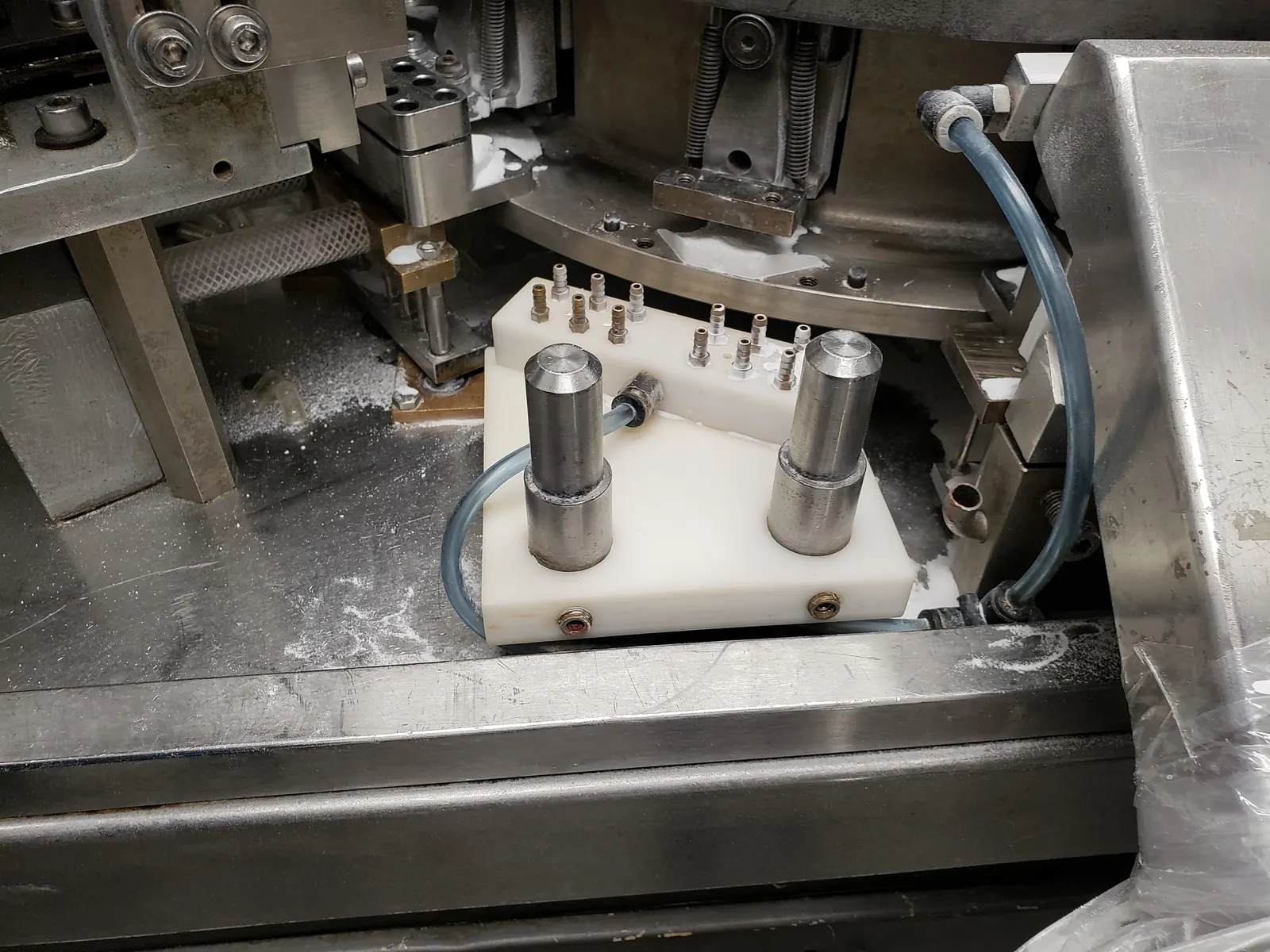 Dosing station inside a capsule filling machine, with a white tooling block, two steel tamping pins and vacuum lines fitted beside the powder-dusted turret.