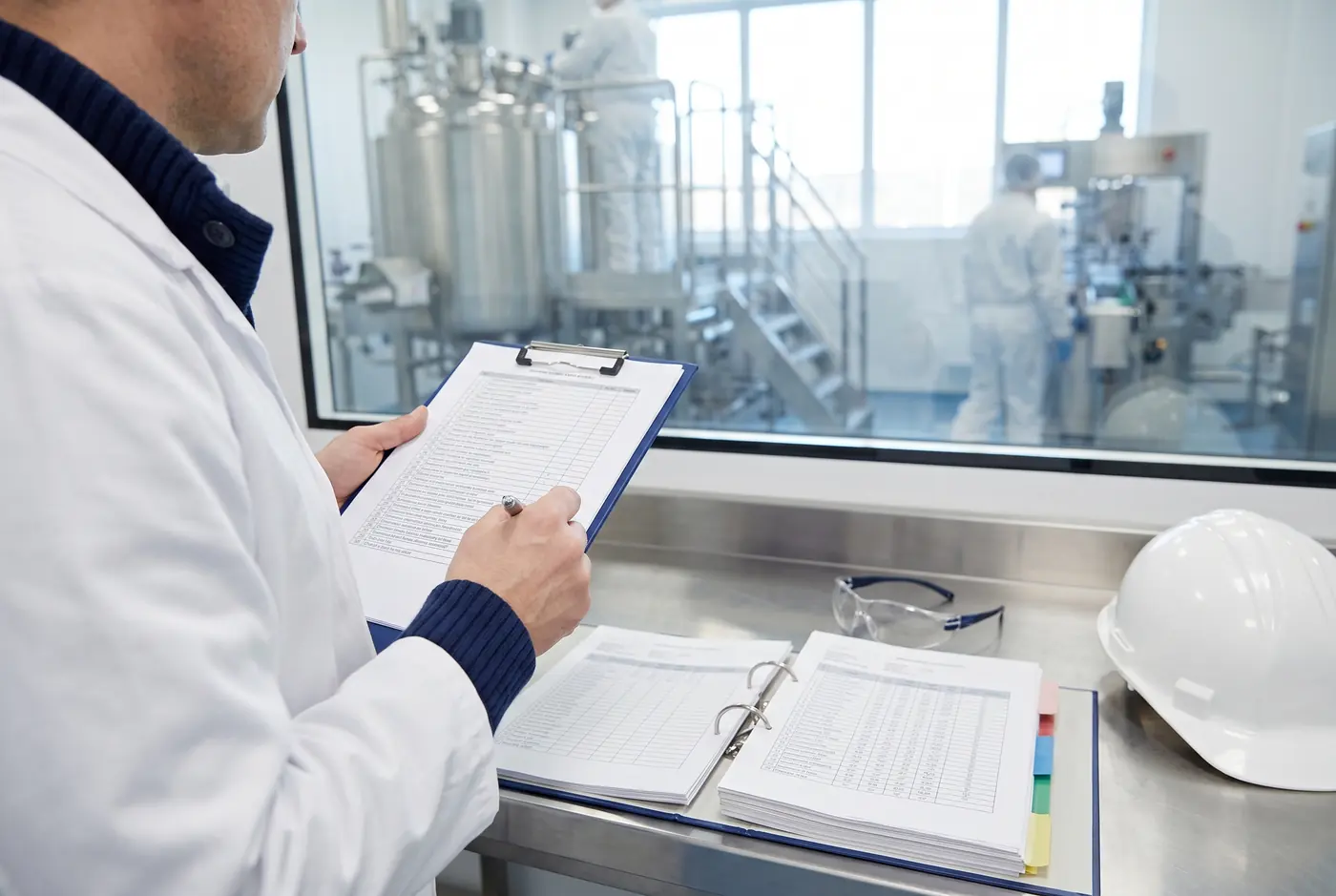 Open batch records and a GMP compliance checklist on a stainless steel desk beside a cleanroom window.